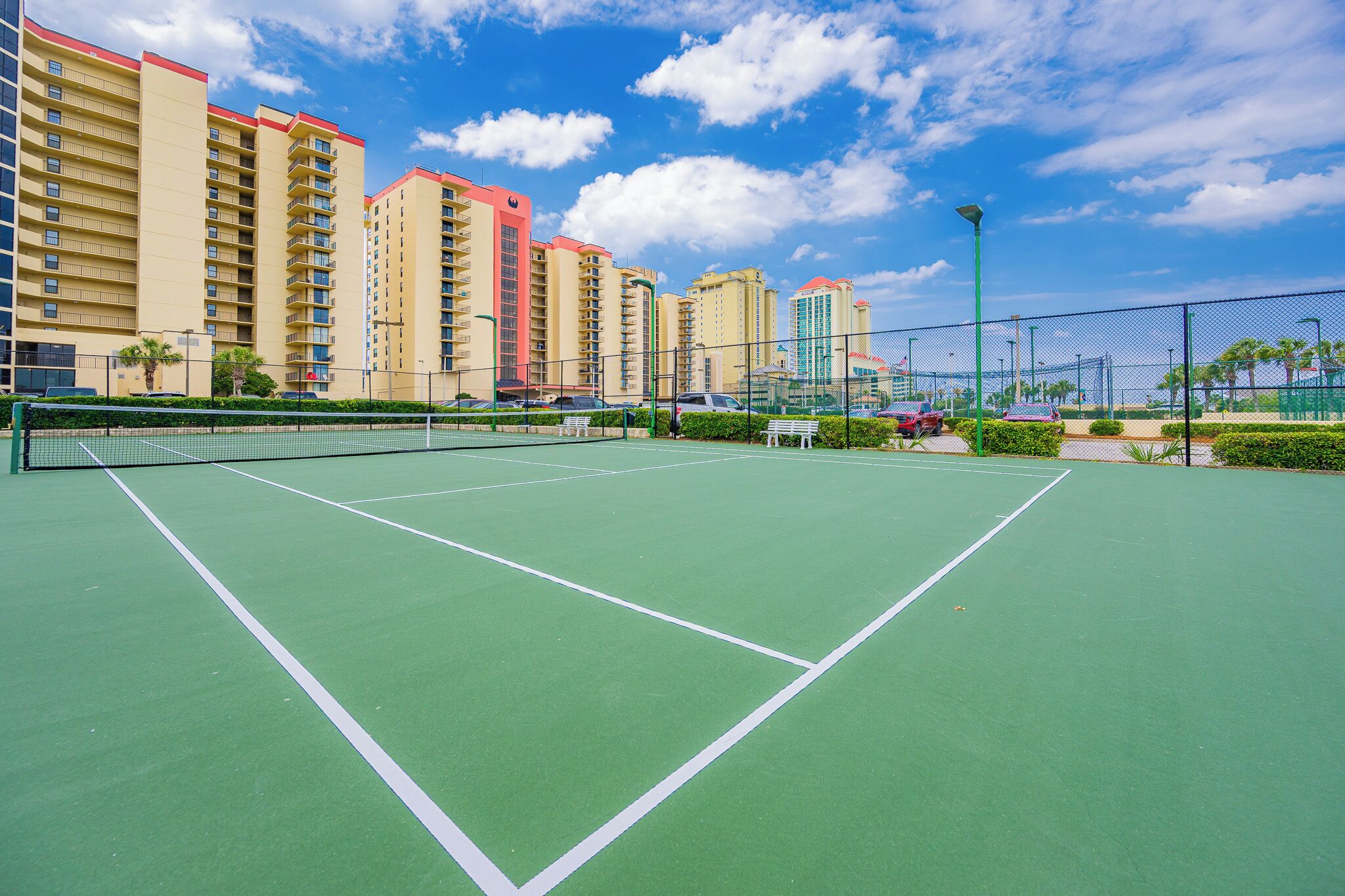 Een tennisbaan met een net, omringd door een hek, naast hoge gebouwen.