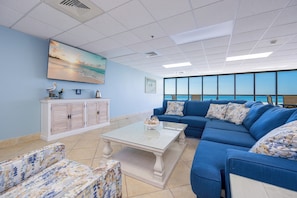A modern lounge with a large window, a blue sofa, a white coffee table, and a cabinet with decorative items. - Two Bedroom @ Phoenix IV (Orange Beach)