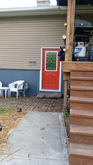 A red door with a decorative glass panel, a small outdoor seating area with white chairs, and a propane tank on a wooden platform.