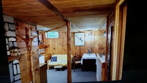 Interior de una cabaña de madera con pared de piedra, una cama, una mesa de comedor y una zona de cocina.