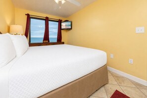 Quarto de hotel com uma cama grande, televisão presa na parede e vista para o mar pela janela.