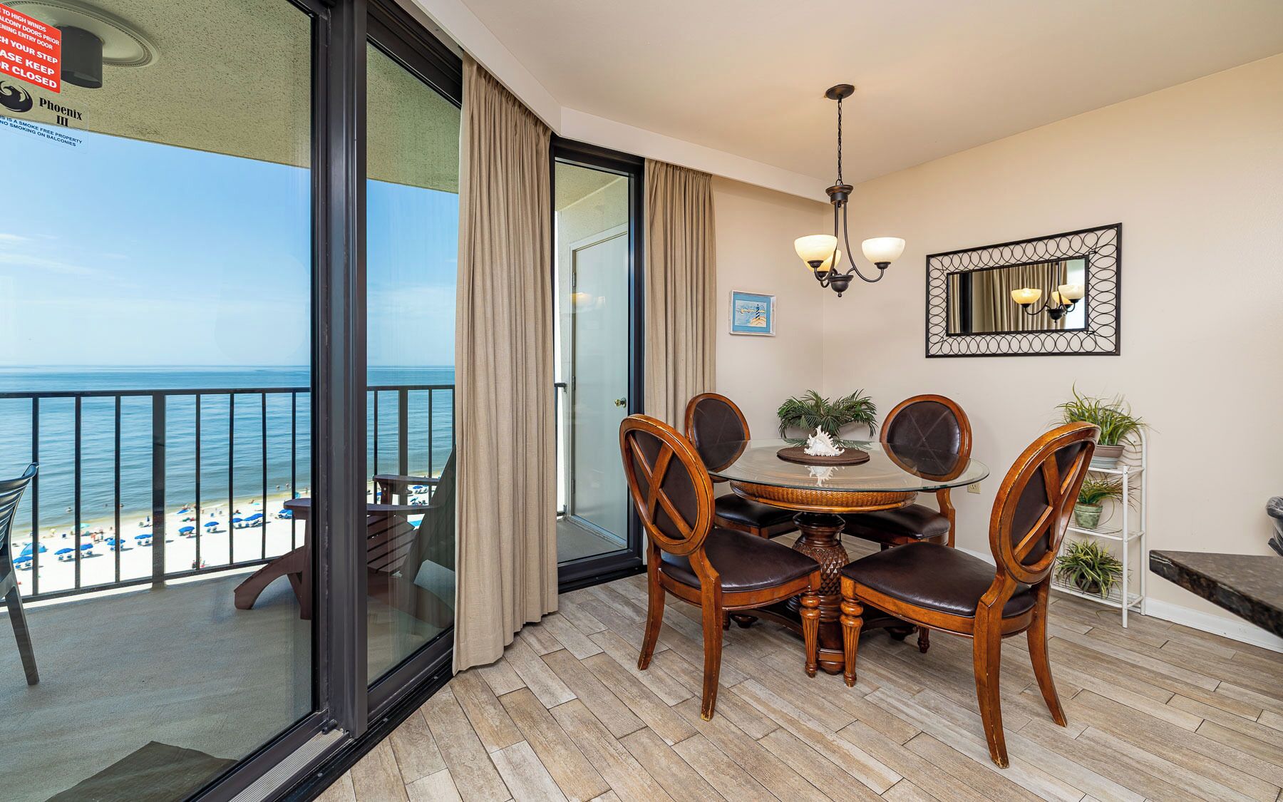 A balcony with a glass door leading to a beach view, a dining table with chairs, and a chandelier.
