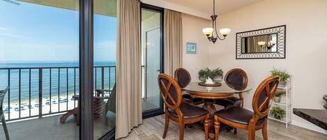 A balcony with a glass door leading to a beach view, a dining table with chairs, and a chandelier.