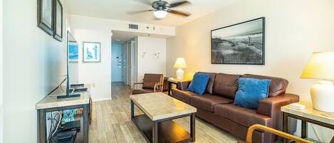 A living room with a brown leather sofa, a coffee table, a desk with a lamp, and a ceiling fan.
