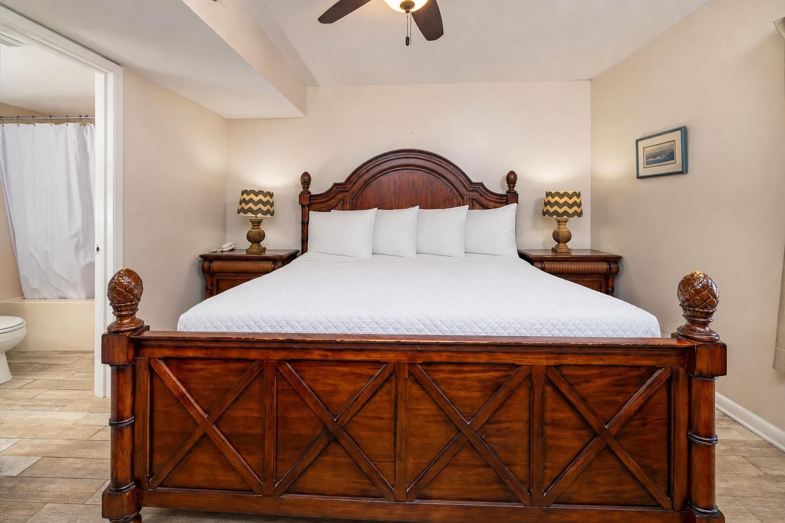 A bedroom with a wooden bed, two bedside tables with lamps, a ceiling fan, and a framed picture on the wall.