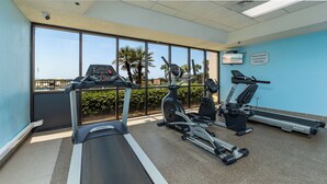 Ein Fitnessraum mit Laufbändern, Crosstrainern und Fahrrädern, mit Blick auf Palmen und einen Pool im Freien.