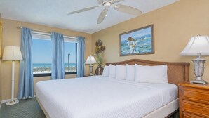 A hotel room with a large bed, a ceiling fan, a painting of a woman on the wall, and a view of the beach through the window.
