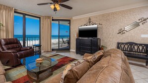 A living room with a leather sofa, a glass coffee table, and a TV on a black cabinet. There is a balcony with a view of the ocean.