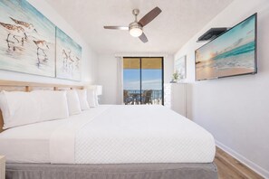 A hotel room with a large bed, a ceiling fan, artwork on the walls, and a view of the ocean through a sliding door. - Reserved Parking with this Two Bedroom at Phoenix 5 (Orange Beach)