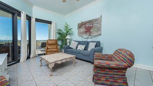 A living room with a blue sofa, a patterned armchair, a wooden coffee table, and a leather armchair. There is a large window with curtains, a plant, and a wall-mounted artwork. - LOW FLOOR Phoenix VII 2BR! (Orange Beach)