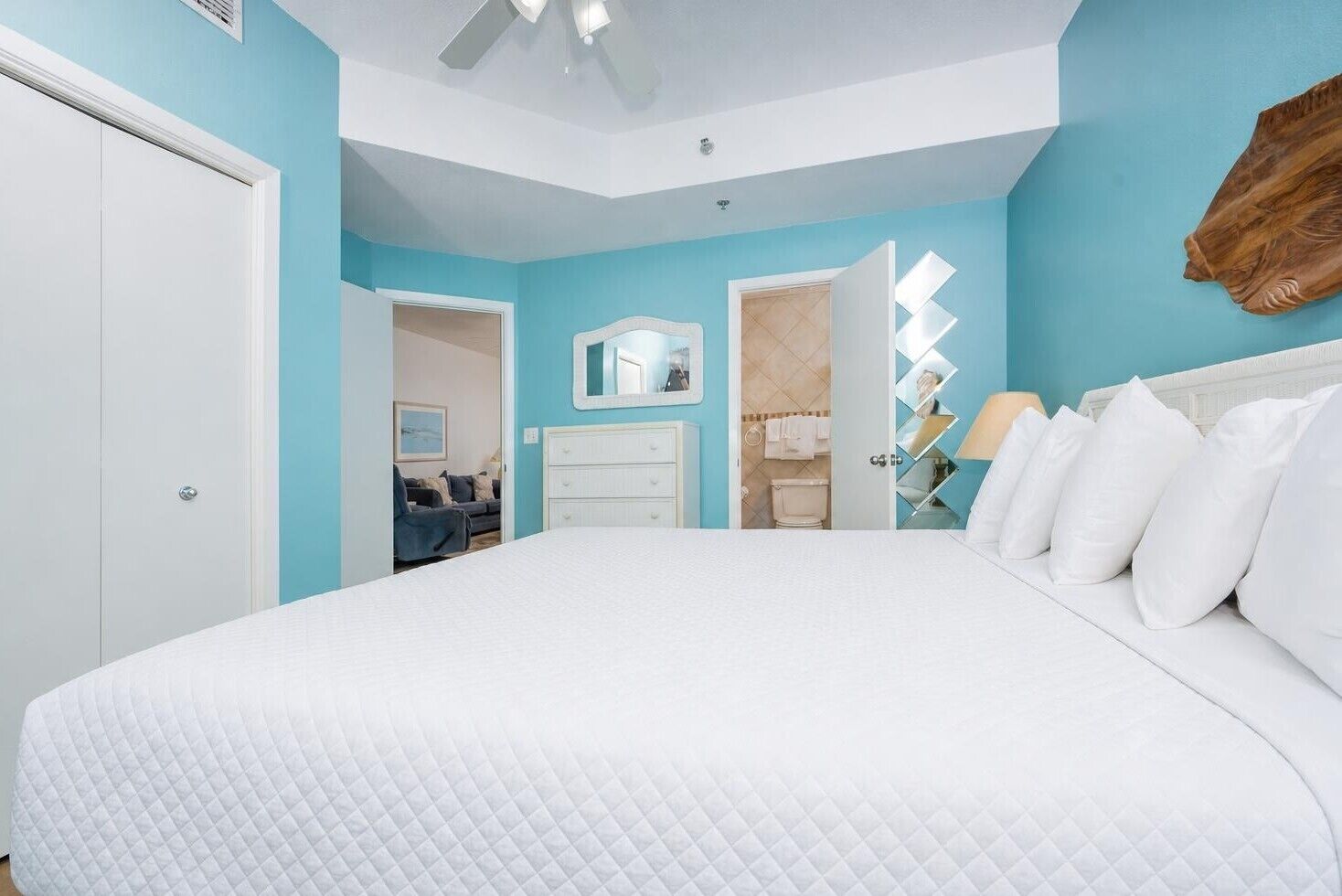 Een slaapkamer met een groot bed, een witte ladenkast, een spiegel en een badkamer zichtbaar door een geopende deur.