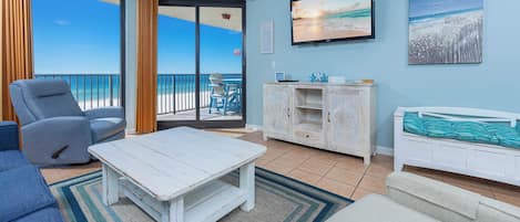 Una habitación con vistas al océano, una mesa de centro blanca, un sillón azul, un armario blanco y un televisor en la pared.