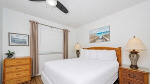 Une chambre à coucher bien rangée, avec un lit, deux tables de chevet, une commode, un ventilateur de plafond, une fenêtre avec des rideaux et un tableau accroché au mur.