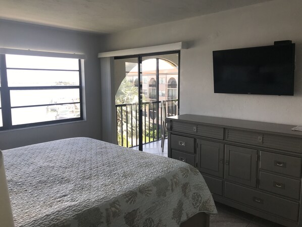 A bedroom with a bed, a dresser, a TV, and a balcony with a view of buildings.