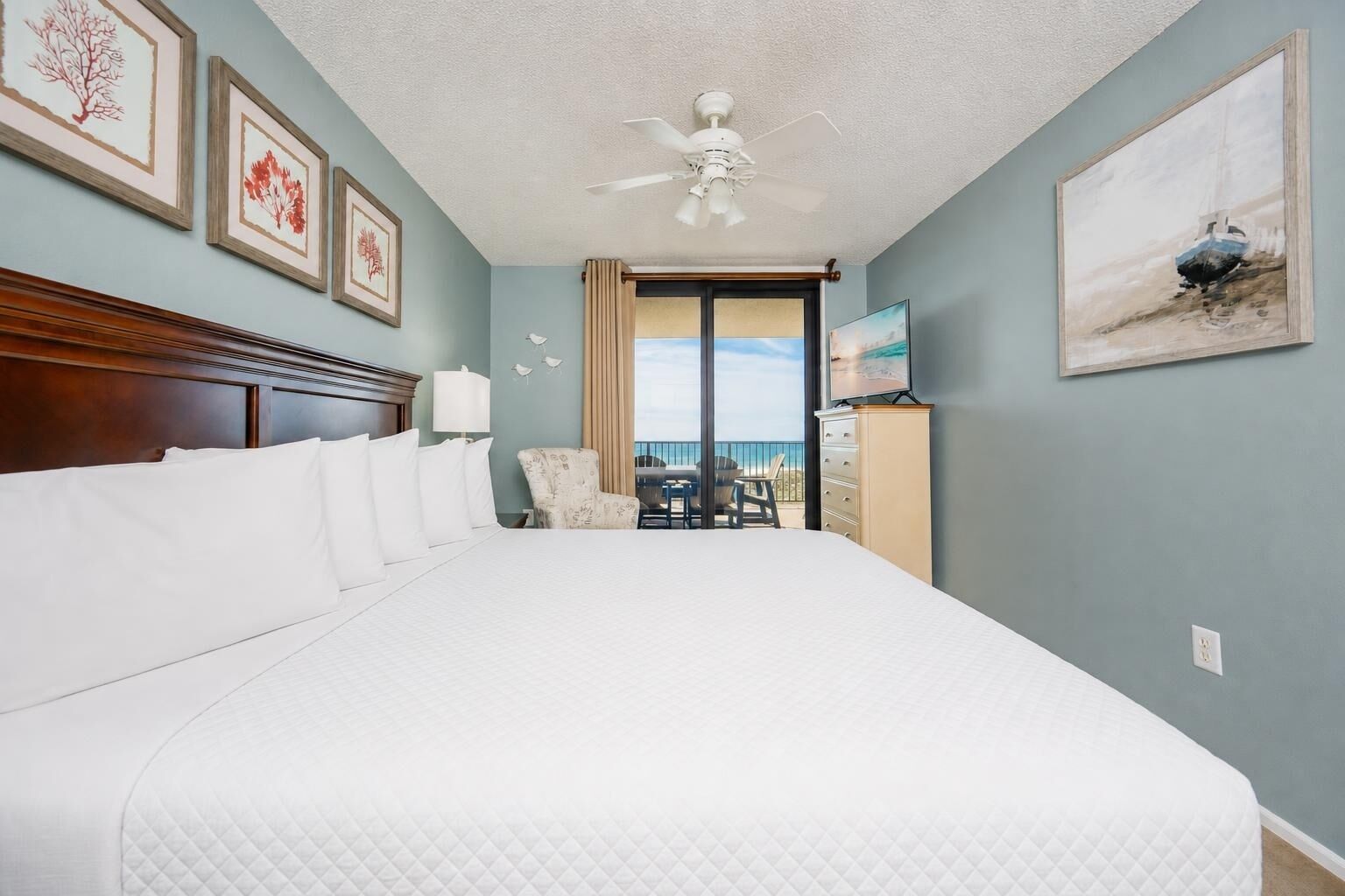Habitación de hotel con una cama grande, ventilador de techo, vista al océano y cuadros en las paredes.