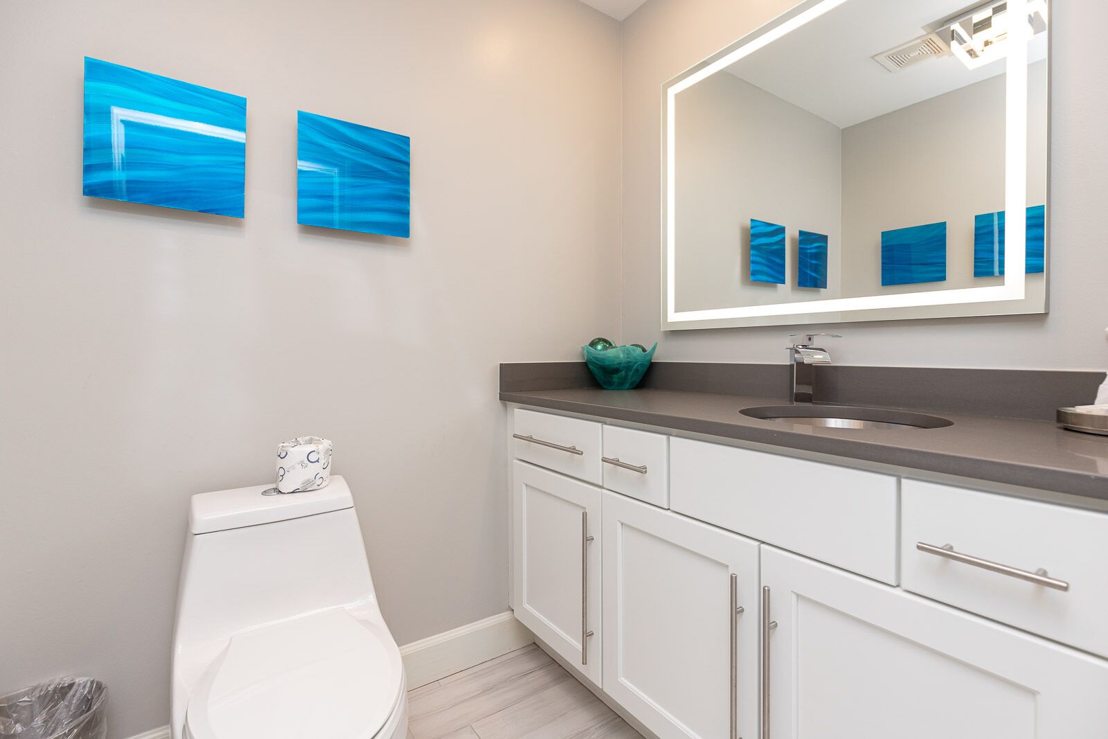 Ein modernes Badezimmer mit Toilette, Waschbecken mit Spiegel darüber und zwei blauen abstrakten Bildern an der Wand.