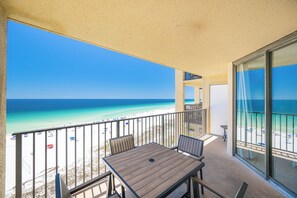 A balcony with a table and chairs, overlooking a beach with a clear view of the ocean and a sandy shore.