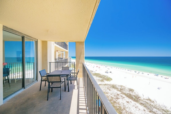 A balcony with a table and chairs, overlooking a beach with clear blue water and a sandy shore.