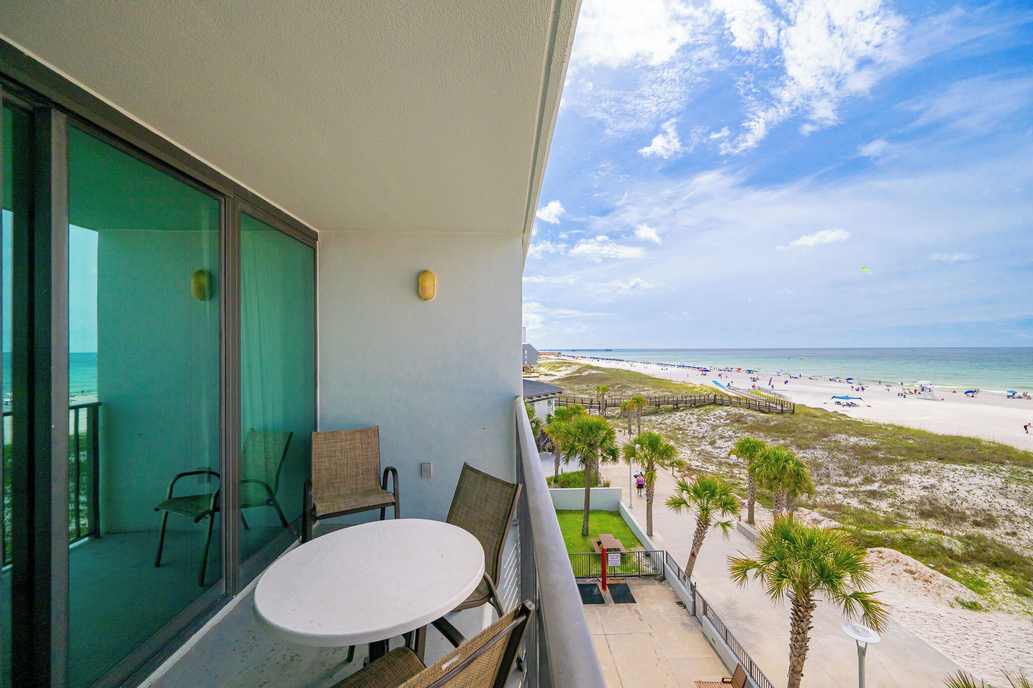 A balcony with a table and chairs, overlooking a beach with palm trees and a boardwalk.