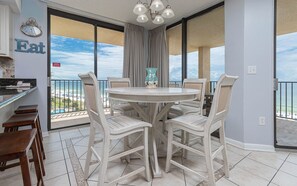 Un comedor con una mesa redonda y sillas, un reloj, y vista a la playa a través de puertas correderas amplias.