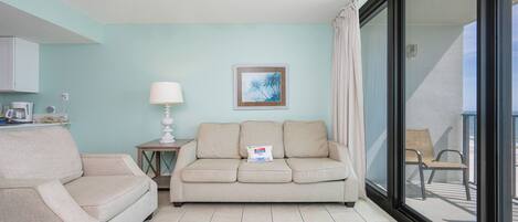 Een woonkamer met een bank, een bijzettafeltje met een lamp, een ingelijst kunstwerk aan de muur en een schuifdeur die leidt naar een buitenruimte.