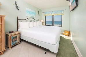 A bedroom with a large bed, a wooden nightstand, a small table, a window with a view of the beach, and a wall-mounted TV.
