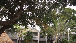 A swimming pool with lounge chairs and a thatched umbrella, surrounded by greenery and a building with balconies.
