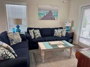 A living room with a dark blue sofa, patterned cushions, a painting of a pier, and a blue vase on a side table.
