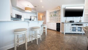 A modern kitchen with white cabinetry, a central island with stools, a built-in oven, and a large flat-screen TV mounted above the island.