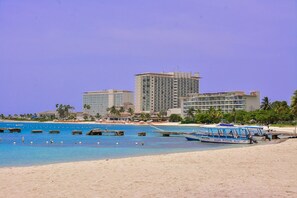 A beach with a large hotel, clear blue water, and a boat docked near the shore. - Coral Keys at Sandcastles (Ocho Rios)