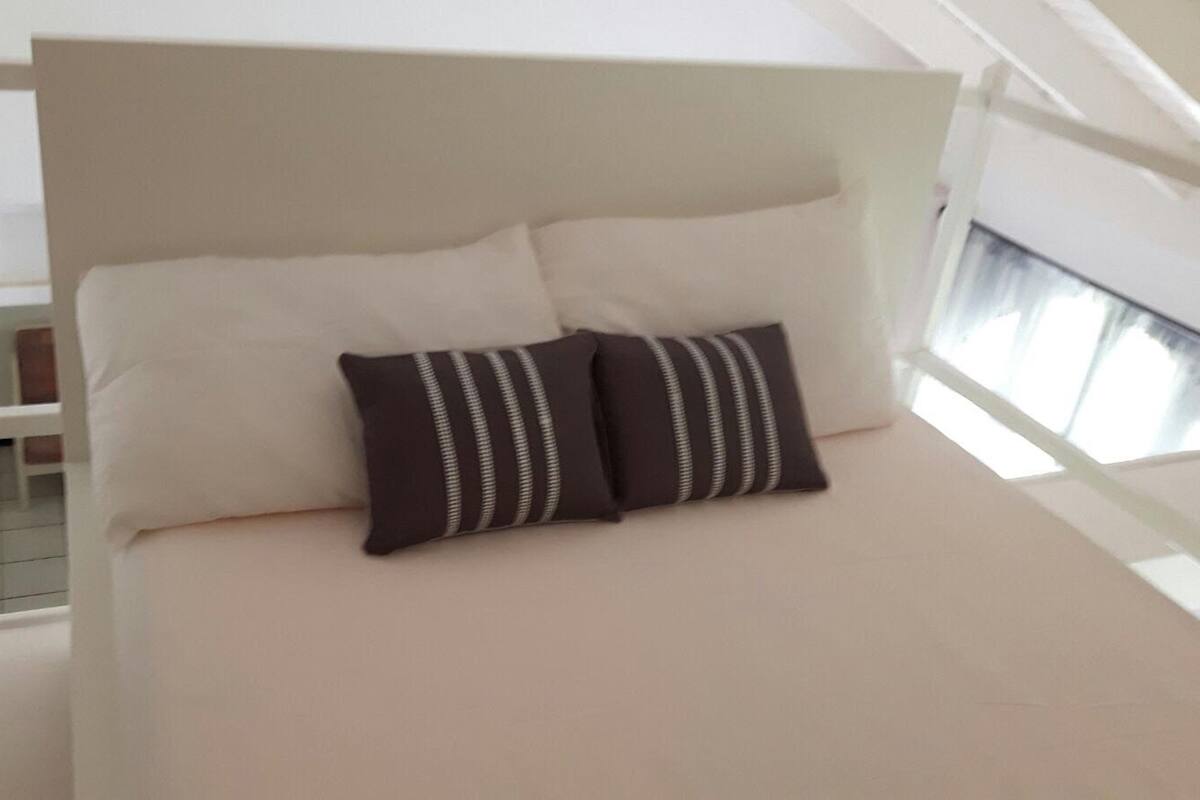 A bed with a white headboard, two pillows, and a light-colored mattress in a room with a window and a white railing.
