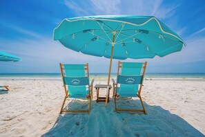Dos sillas de playa con una mesa bajo un paraguas grande en una playa arenosa.