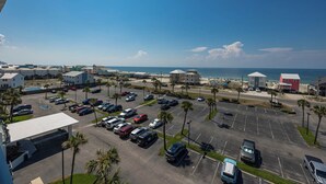 A parking lot with multiple cars, a beach in the background, and several buildings including a red one.