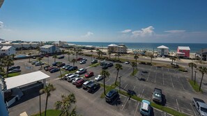 A parking lot with multiple cars, a beach in the background, and several buildings including a red one.