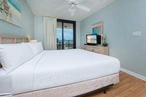 A hotel room with a bed, a television, a ceiling fan, and a view of the beach through a sliding door.