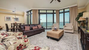 A spacious living room with a large brown sofa, two beige armchairs, a coffee table, and a dining area with a table and chairs. - The Oasis at Orange Beach, formerly known as Phoenix West II, Unit 503 (Orange Beach)