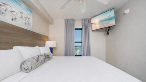 A hotel room with a large bed, a flat-screen TV, a ceiling fan, and a view of the outdoors through a window.