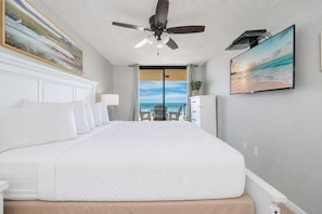 A hotel room with a large bed, a ceiling fan, a TV, a painting, and a view of the ocean.