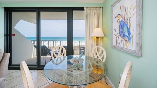 Una mesa de comedor con cubierta de vidrio, sillas blancas, vista al mar a través de un vidrio corredero y un cuadro enmarcado de un pájaro en la pared.