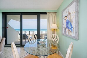 Una mesa de comedor con cubierta de vidrio, sillas blancas, vista al mar a través de un vidrio corredero y un cuadro enmarcado de un pájaro en la pared.