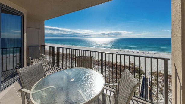 A balcony with a glass table and chairs, overlooking a beach with a clear view of the ocean.