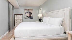 Una cama bien hecha en una habitación con pared azul claro, un cuadro de un barco, un tocador blanco y una lámpara de noche.