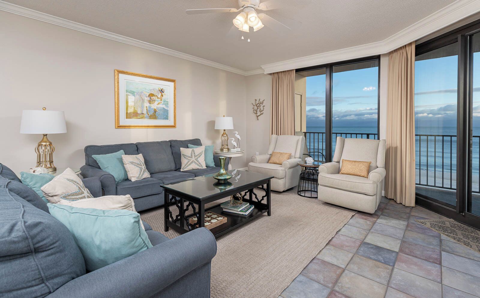 A spacious living room with a grey sofa, a coffee table, and a beige armchair. A large window offers a view of the ocean.