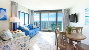 A room with a blue sofa, a wooden dining table, and chairs, a TV mounted on the wall, and a balcony with a view of the sea.