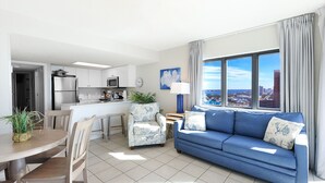 A modern living room with a blue sofa, a dining table with chairs, a kitchen area with white cabinets, and a large window offering a city view.