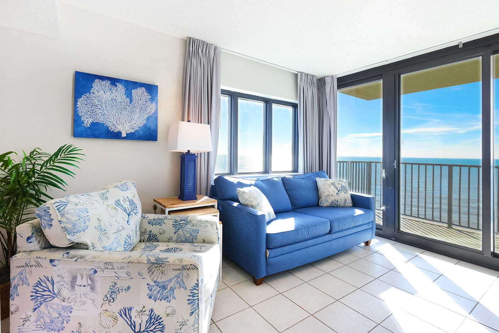 A living room with a blue sofa, a patterned armchair, a table with a lamp, and a large window with a view of the sea.