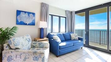 A living room with a blue sofa, a patterned armchair, a table with a lamp, and a large window with a view of the sea.