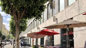 Un café al aire libre con mesas y sillas, un árbol y un edificio con amplios ventanales.