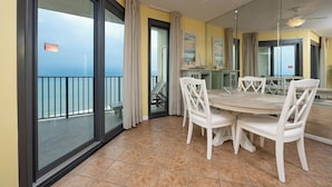 Un comedor con una mesa redonda y sillas blancas, un balcón con vistas al océano y una cocina visible a través de una puerta de cristal.