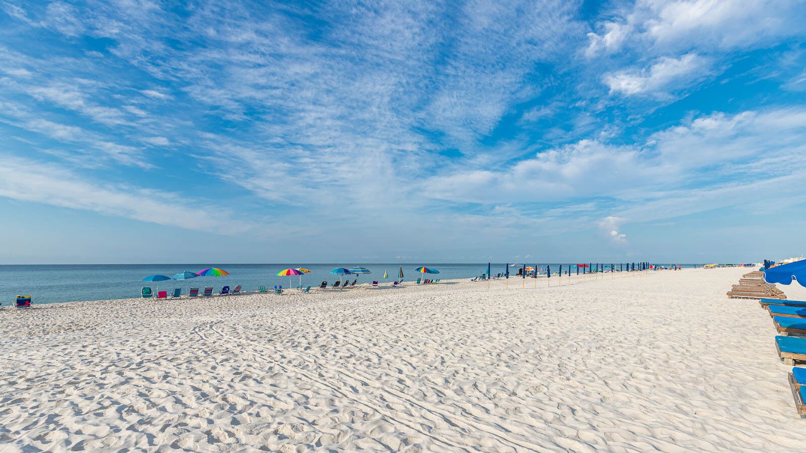 Une vaste plage de sable fin, bordée de rangées de chaises longues et de parasols, une jetée s’avançant dans l’eau et un ciel bleu azur.